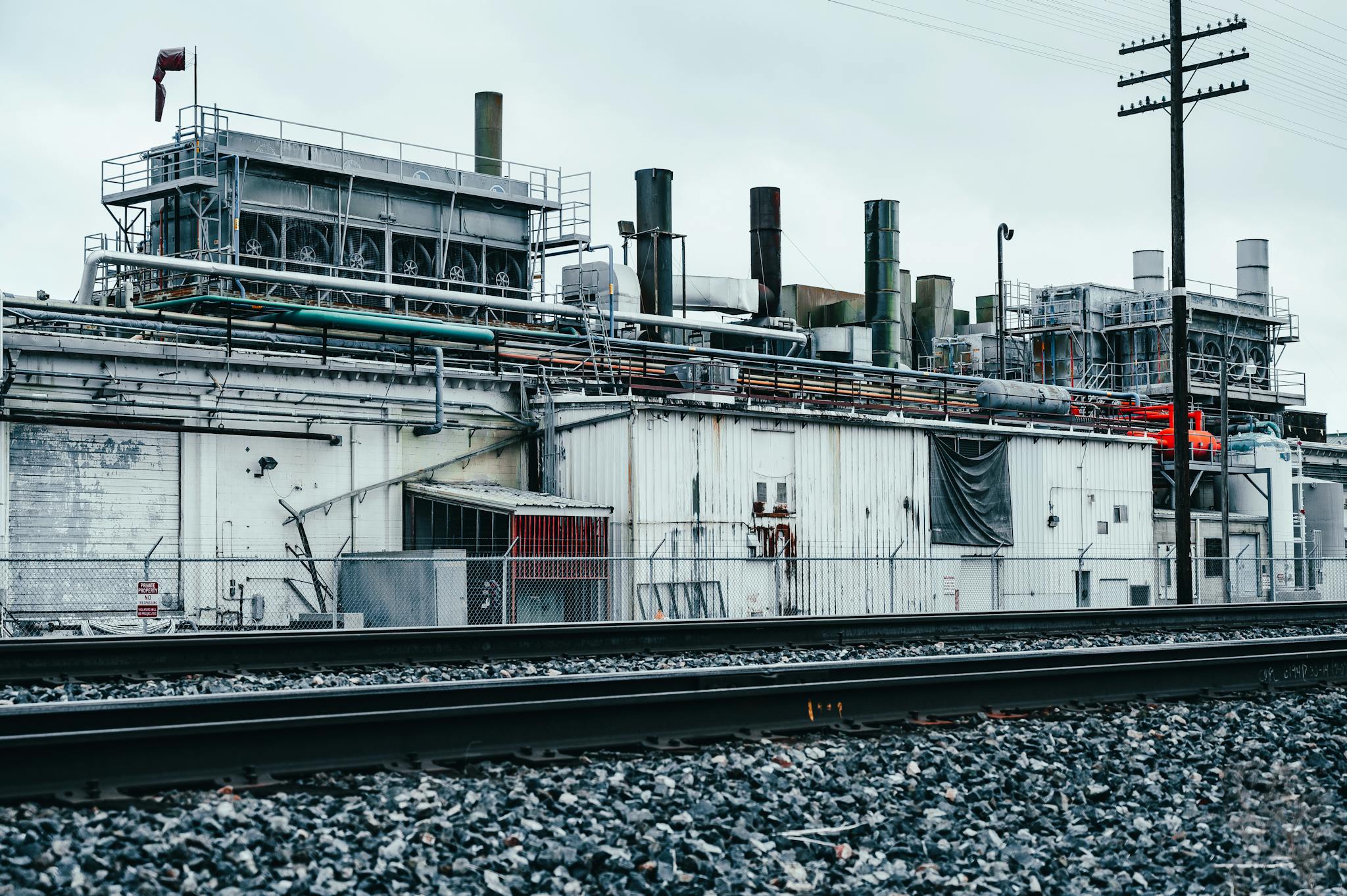 A weathered factory building with visible pipelines and railway tracks under an overcast sky.